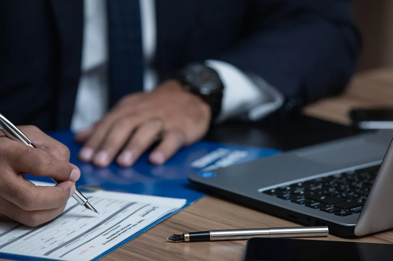Una persona firma un documento junto a una laptop, sobre un escritorio de madera